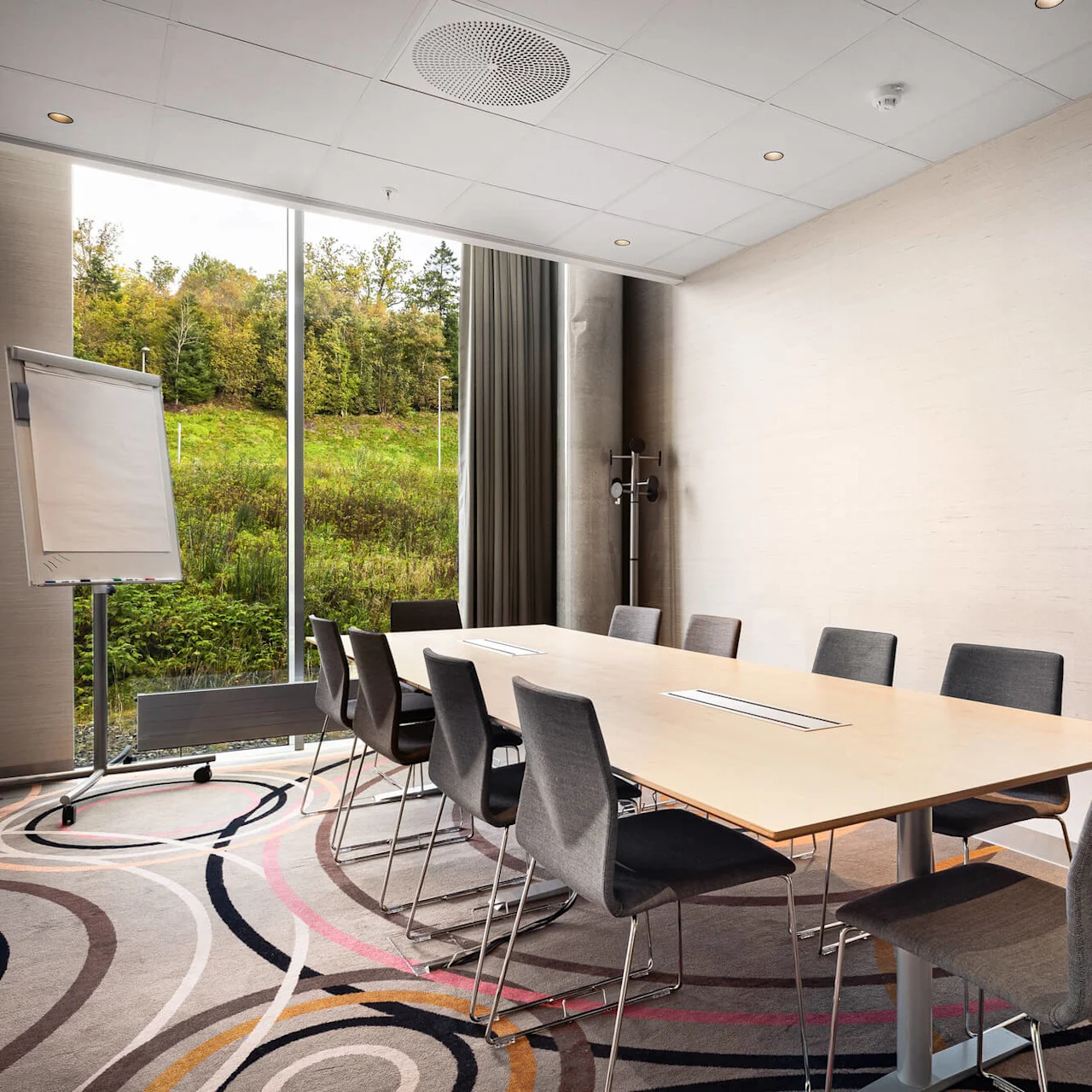 A conference room features a long wooden table surrounded by chairs. A flip chart stands nearby, with a window overlooking a grassy slope with trees outside.