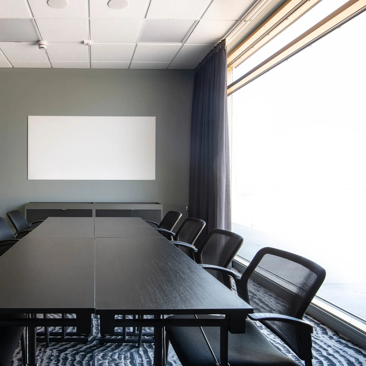 A long black conference table surrounded by black chairs sits in a meeting room with a whiteboard on a gray wall. Large windows with curtains provide natural light.