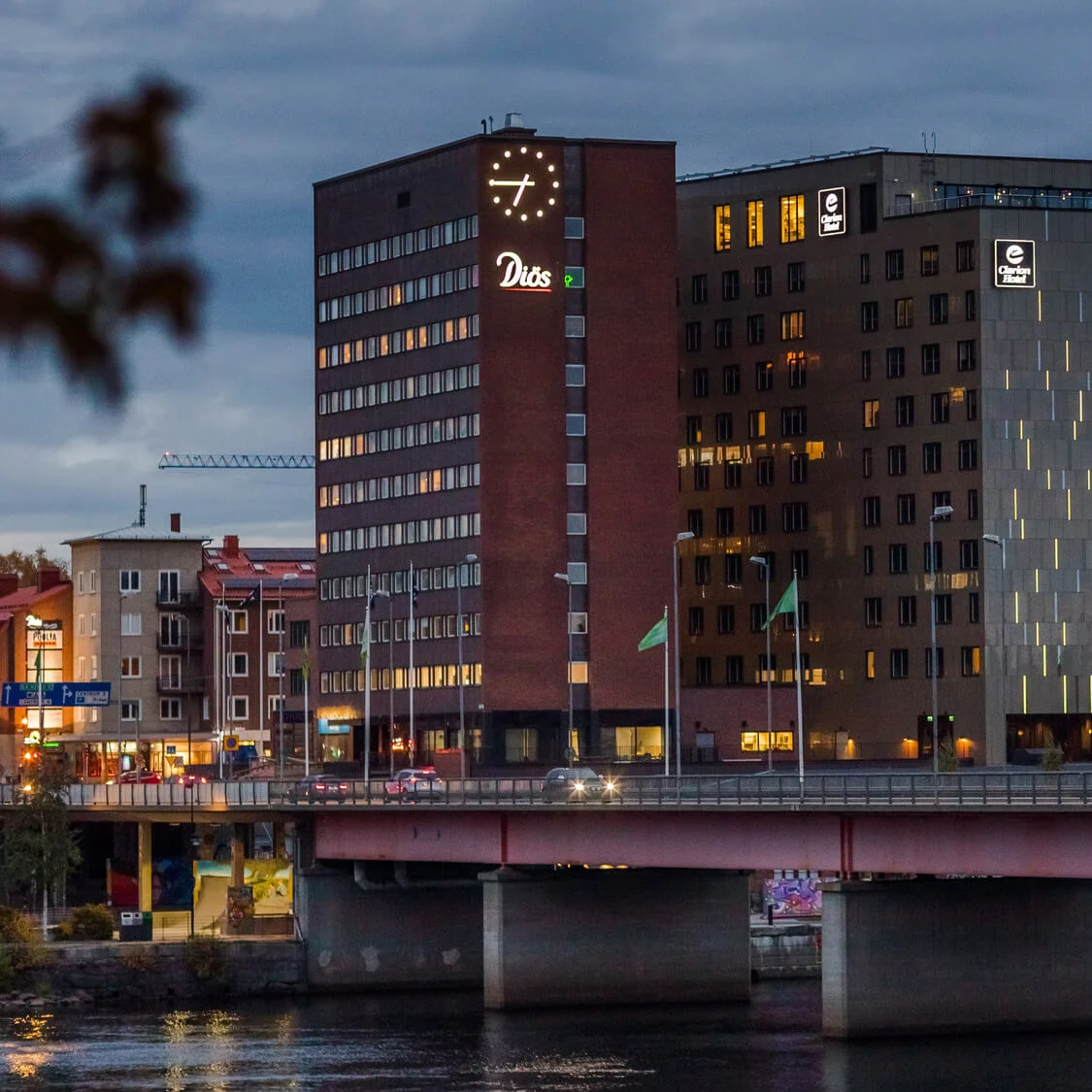 En bro strækker sig over en flod i et byområde, med høje bygninger og oplyste hoteller i baggrunden. Nogle bygningsskilte er tændt, inklusive "Dios" og "Clarion Hotel."
