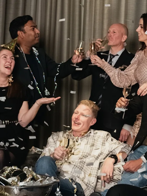 En grupp människor firar, ler och håller champagneglas, omgiven av konfetti. De är klädda i festkläder med en gardin som bakgrund.