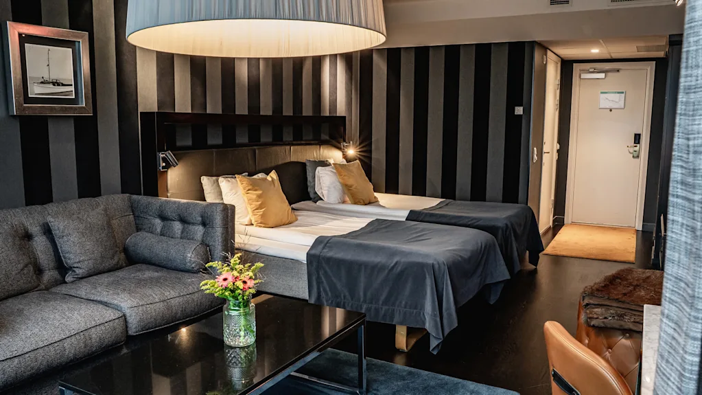Ett modernt hotellrum innehåller två sängar med mörka sängkläder, en grå soffa och ett svart soffbord med blommor. En stor lampa hänger ovanför; randig tapet och en stängd dörr syns.