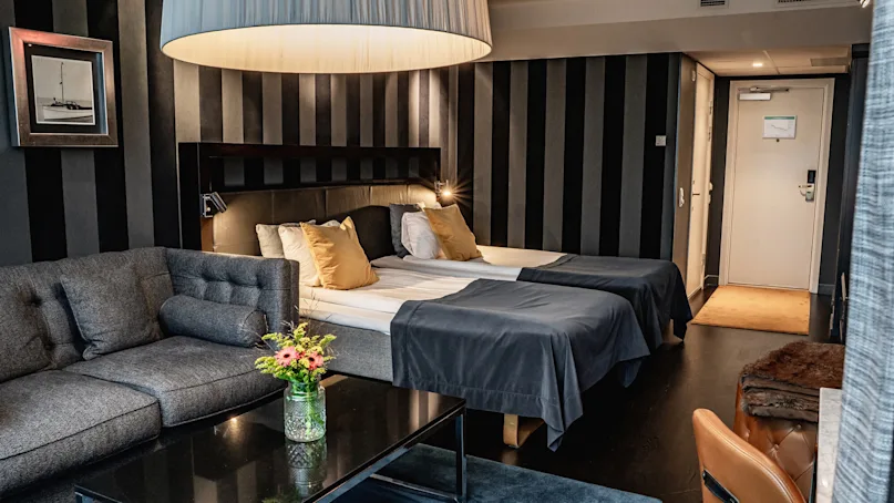 A modern hotel room contains two beds with dark linens, a gray sofa, and a black coffee table with flowers. A large lamp hangs above; striped wallpaper and a closed door are visible.