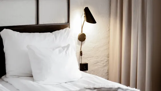 A neatly made bed with white bedding and beige curtains in a softly lit bedroom, a wall-mounted lamp turned on.