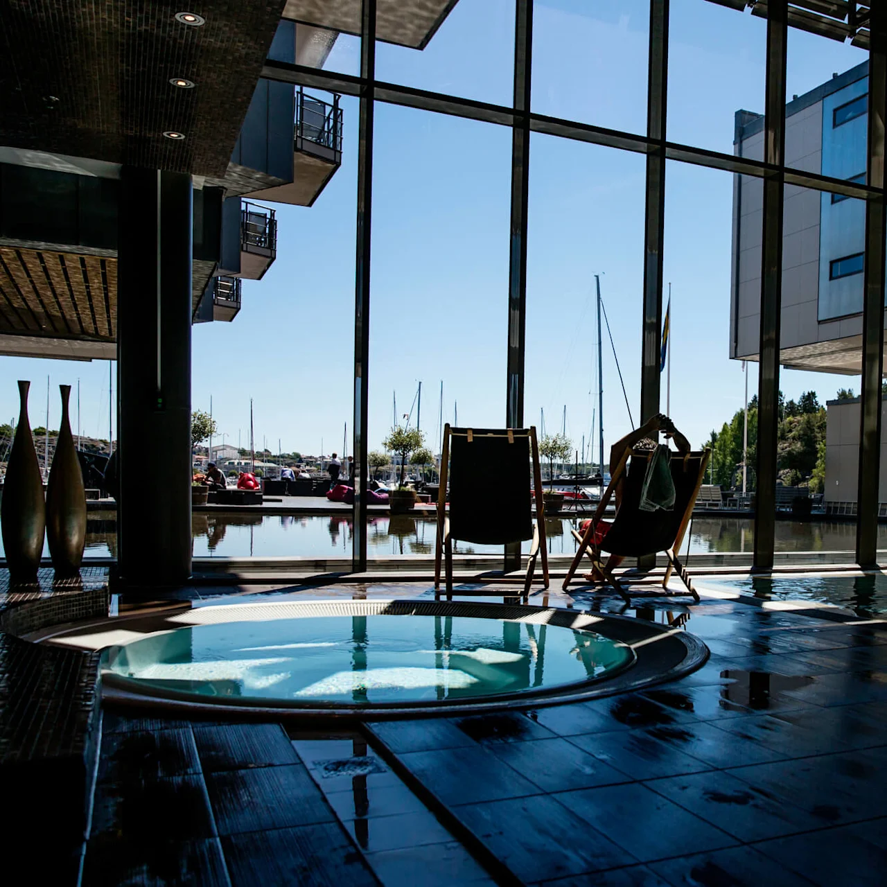 Et boblebad står stille i et moderne interiør, ved siden av to dekkstoler vendt mot store vinduer med utsikt over marinaen, utenfor solbelyst blå himmel og båter til kai.