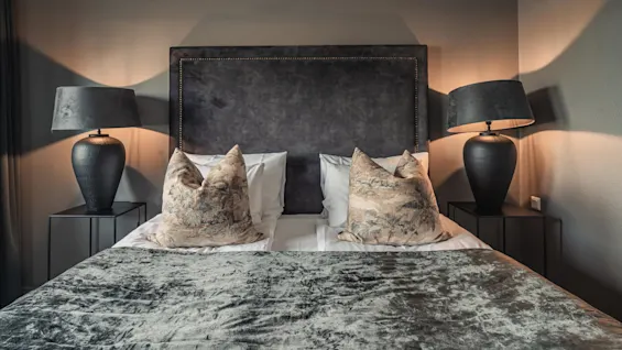 A bed with patterned pillows and a dark headboard is flanked by two nightstands with large black lamps, set in a softly lit bedroom.
