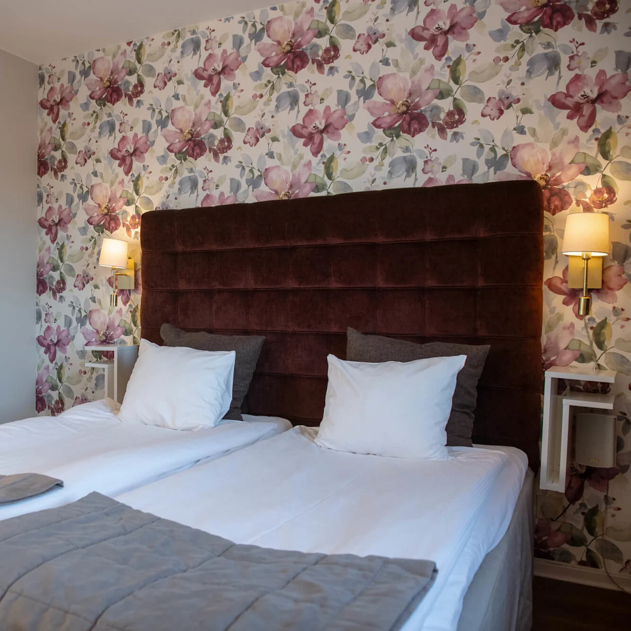 A neatly arranged bed sits against a floral wallpapered wall, with brown headboard and two framed floral artworks beside it, in a softly-lit bedroom setting.