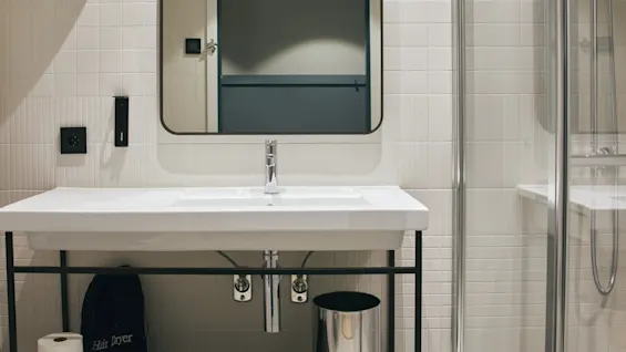 Ett modernt hotellbadrum på Stopover Hotel Falkenberg med handfat, spegel och dusch. En väska för en Hair Dryer förvaras under handfatet.
