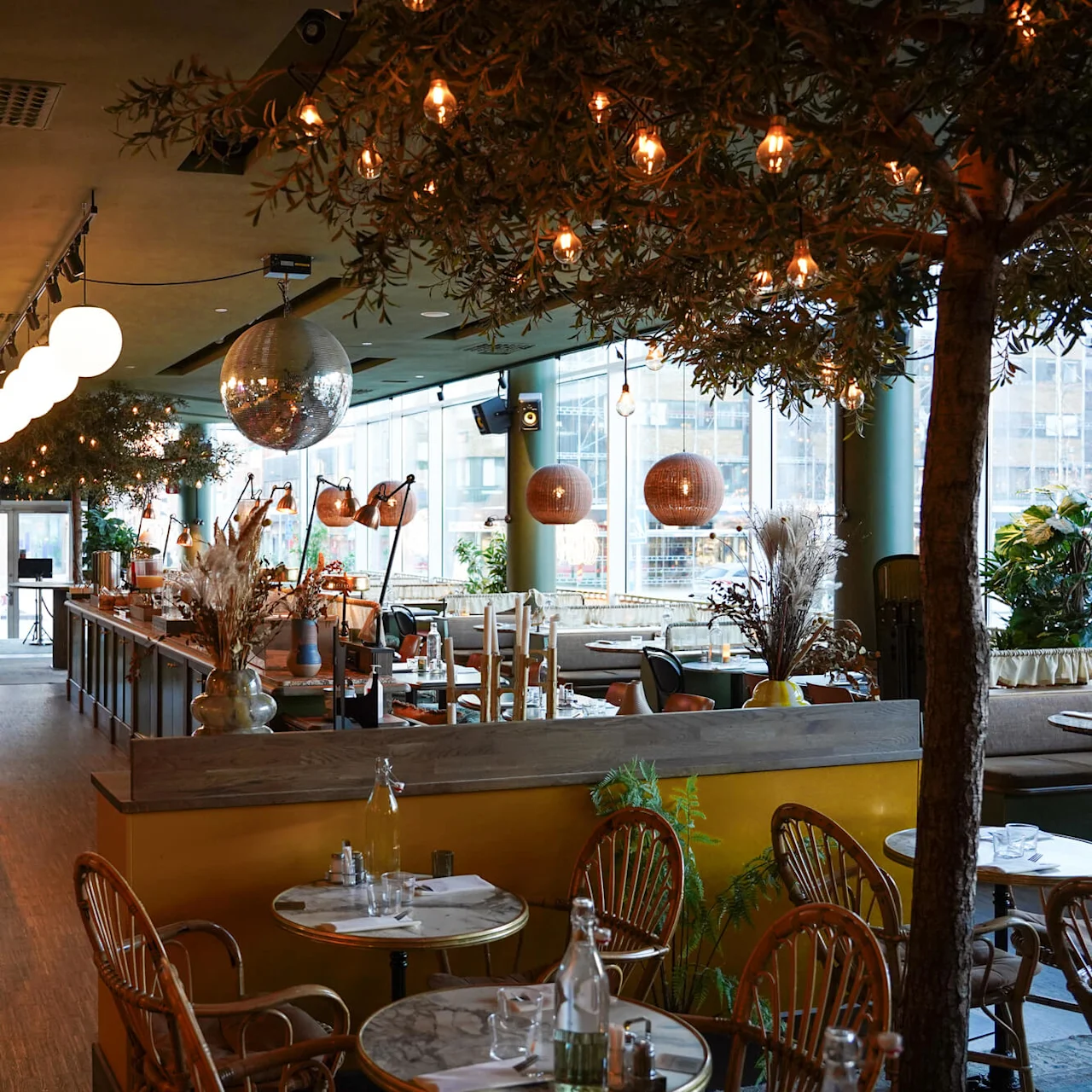 En varmt belyst restaurantinteriør med eklektisk dekoration, møbler arrangeret til spisning, og træer som øger den hyggelige stemning.