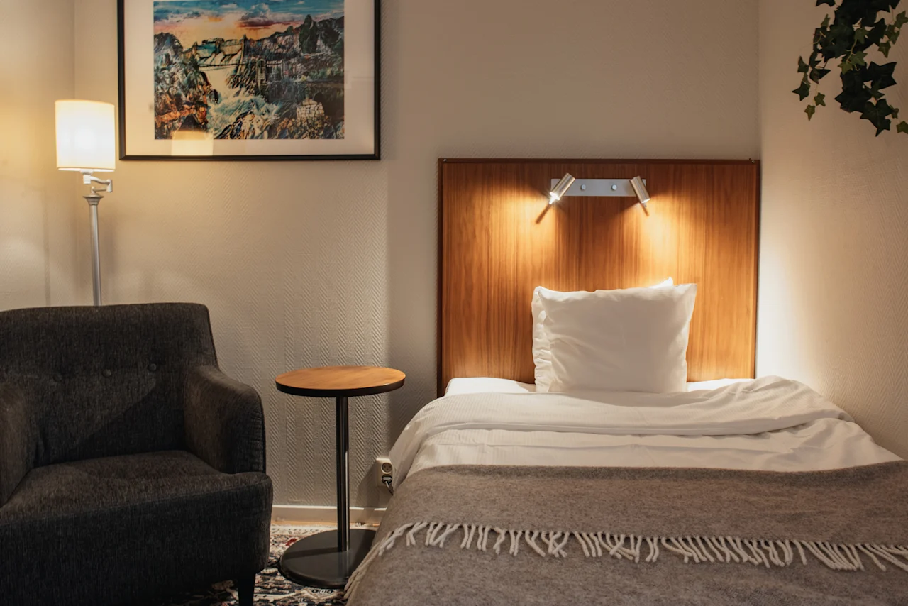 Home Hotel Kung Oscar – Compact Cabin – A cozy hotel room with a single bed, an armchair, and a side table, illuminated by soft lighting.
