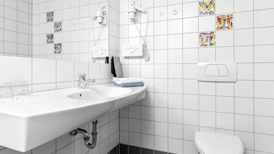 A white ceramic sink with a silver faucet holds folded towels in a tiled bathroom. Glasses sit on the countertop. A wall-mounted hairdryer and toilet are visible nearby.