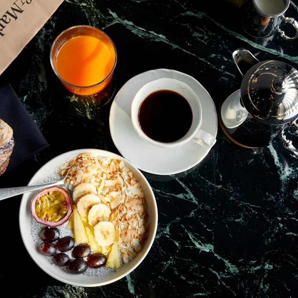 En frokostoppsett på et mørkt marmorbrett inkluderer en bolle med skivede bananer, kokos, druer og pasjonsfrukt. Ved siden av er det kaffe, appelsinjuice, et bakverk og en avis.