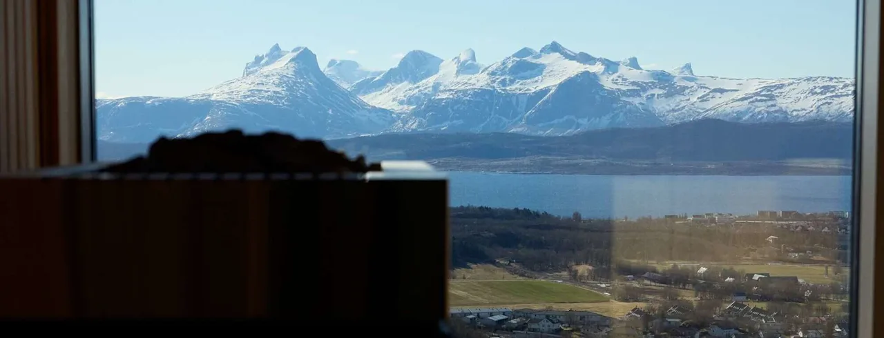 En saunaovn er silhouet i forgrunden, med sneklædte bjerge synlige gennem et stort vindue, med udsigt over en by under en klarblå himmel.