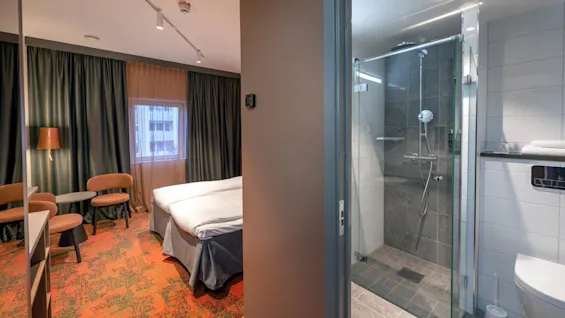 A hotel room features a neatly made bed beside a window with dark curtains, orange carpet, and two chairs. Adjacent is a modern bathroom with a glass-enclosed shower.
