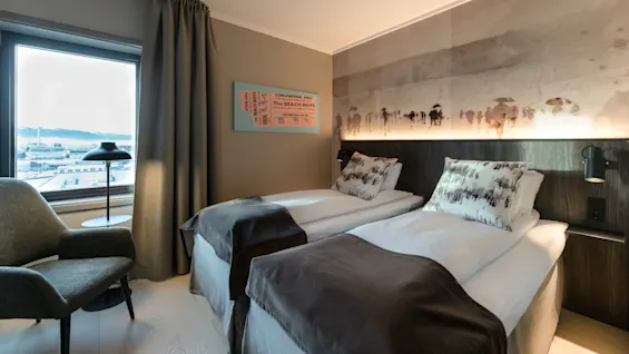 To senger med hvitt sengetøy og mørke tepper i et hotellrom, dekorert med abstrakt kunstverk. Et vindu viser en byutsikt. Tekst: "The BEATLES in SWEDEN."