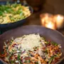 A colorful salad with shredded vegetables and sprouts sits in a brown bowl. Another bowl with pasta and greens in the background. The setting is warmly lit by candles.