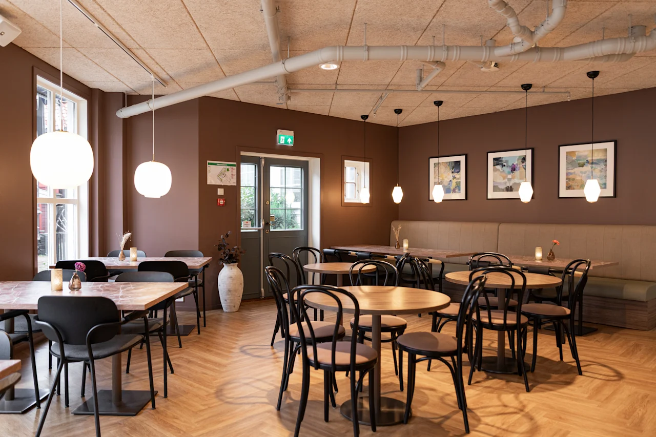 A cozy cafe interior with wooden floors, brown walls, and various tables and chairs, illuminated by hanging lights.