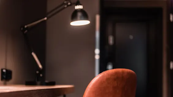 A brown leather chair sits by a wooden desk, illuminated by a black desk lamp, in a dimly lit room with dark walls.