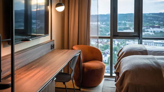 Et trebord med en stol er ved siden av en veggmontert TV i et rom med store vinduer med utsikt over et bymiljø. Nærheten er to senger og en oransje lenestol.