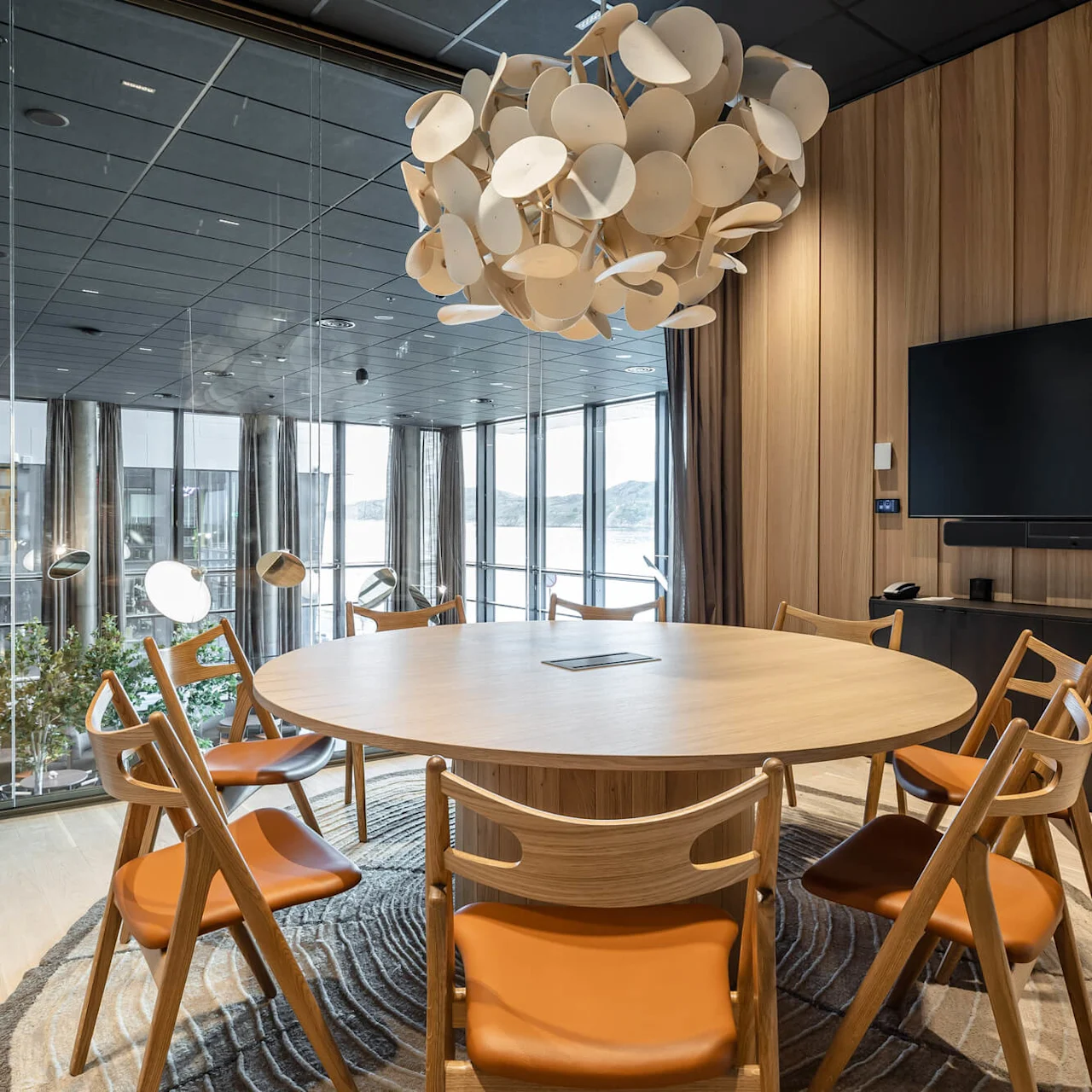 Round wooden table surrounded by chairs with leather seats; hanging modern lamp above. Set in a glass-walled room with a view of trees and mountains outside.