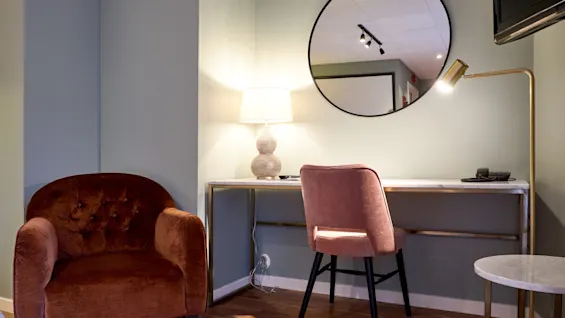 A plush chair and desk with a lamp, beneath a round mirror, in a softly lit room.