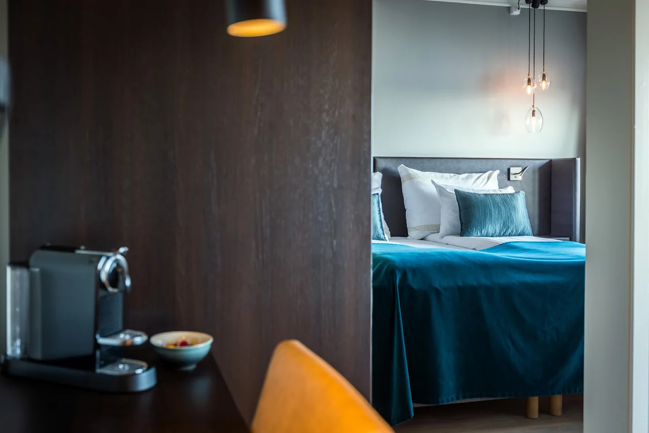 Bed with teal blanket and pillows, softly lit by hanging lamps, adjacent to a wooden desk with a coffee machine and a bowl; modern bedroom setting.