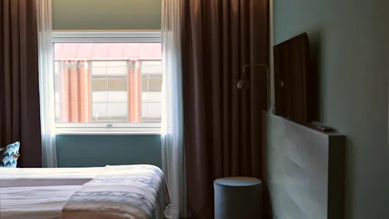 En pænt redt seng vender mod et vægmonteret tv. Mørke gardiner indrammer et vindue med byudsigt. En rund skammel står i nærheden, hvilket fremhæver rummets moderne stil.