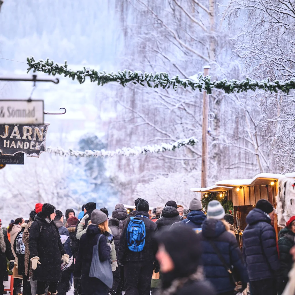 People enjoying a snowy Christmas market in Östersund with festive decorations and market stalls. Signs read Trall & Sonnad and Jarn Handel.