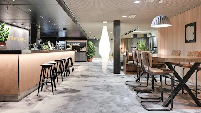 En moderne hotellsalong har en elegant bar med høye krakker, et langt felles bord og stilig dekor. Planter gir grønt miljø. Tekst på veggen: "COMFORT HOTEL XPRESS."