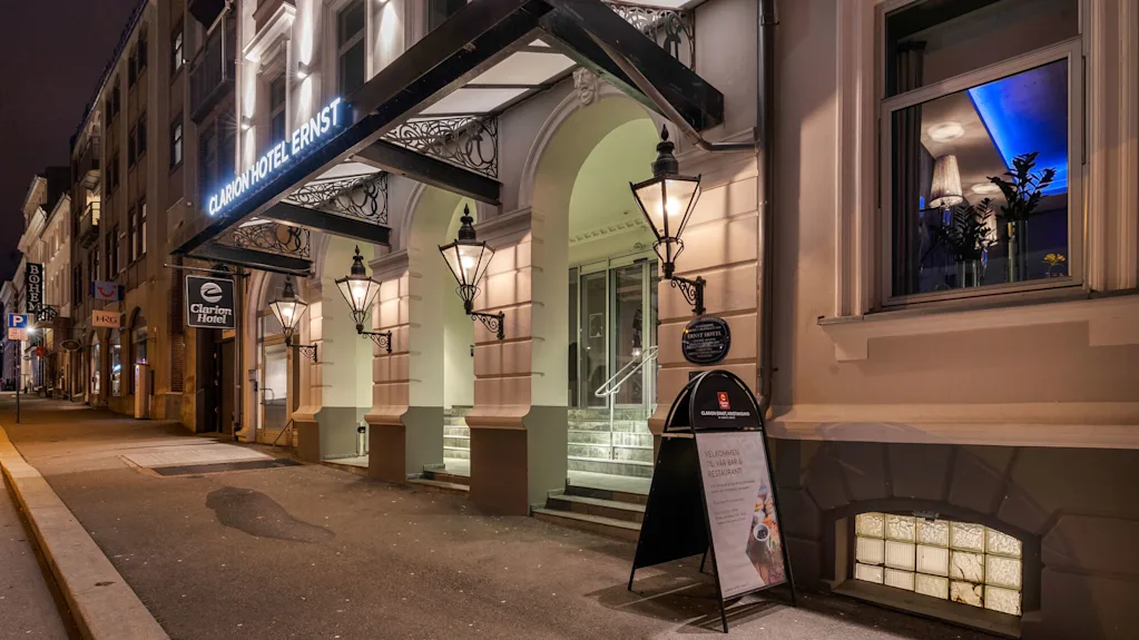 Clarion Hotels ingång upplyst på natten, med arkitektoniska detaljer och skyltar. I närheten står en gatupratare med texten: "Velkommen til vår bar & restaurant!" Gatuvy inkluderar intilliggande byggnader.