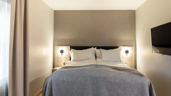Et koselig hotellrom med en pent oppredd seng, nattbordslamper og en veggmontert TV, klart for et behagelig opphold.