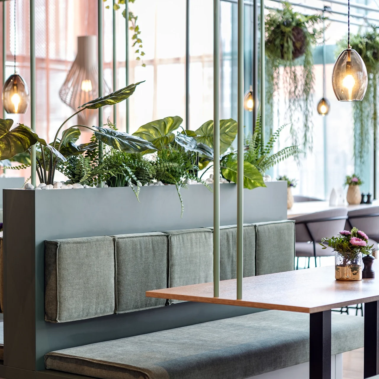 Gröna vadderade bås omger träbord, dekorerade med frodiga växter och hängande pendellampor i en modern, ljus matsal.