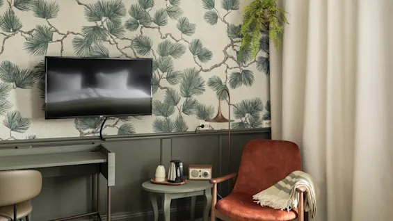 Kutsuva hotellihuone, jossa on televisio, toimiva työpöytäalue ja muhkea nojatuoli rentoutumiseen, koristeltu luontoaiheisella tapetilla.