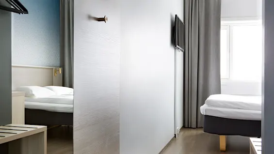 A modern, minimalistic hotel room split by an open door; two twin beds, soft lighting, wall-mounted TV, and curtains in a cozy ambiance.
