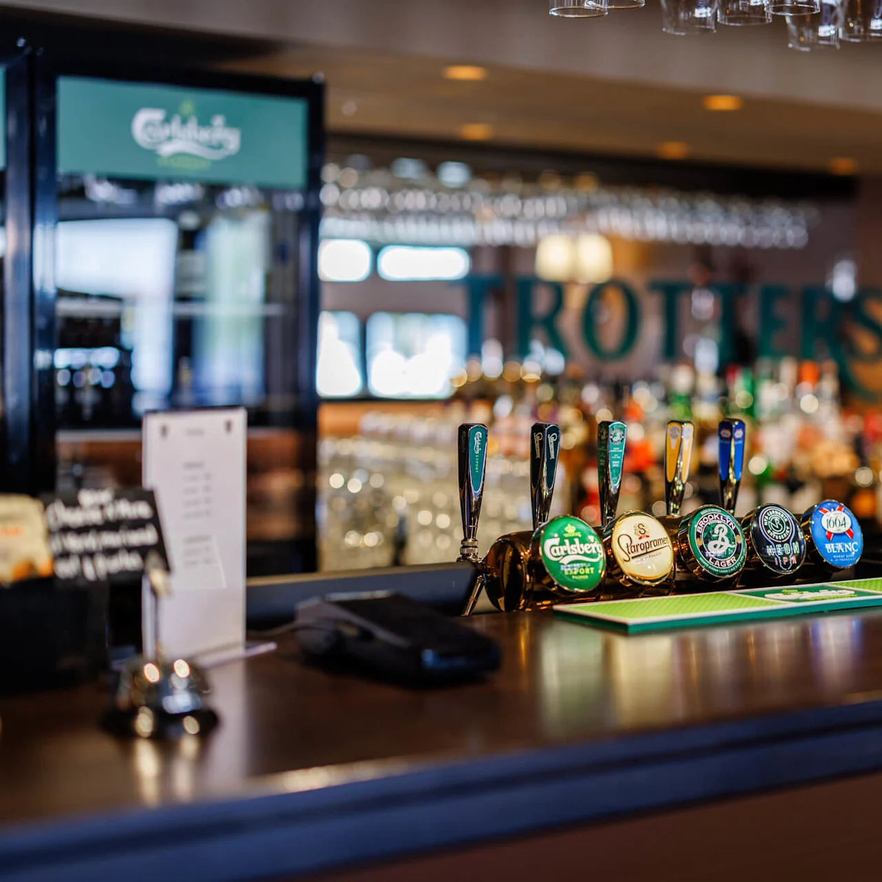 Ölkranar med olika märkesetiketter är uppradade på en bardisk i trä i en pub. Ovan hänger glas upp och ner. I bakgrunden syns en suddig bardisk. Text: "Guinness."