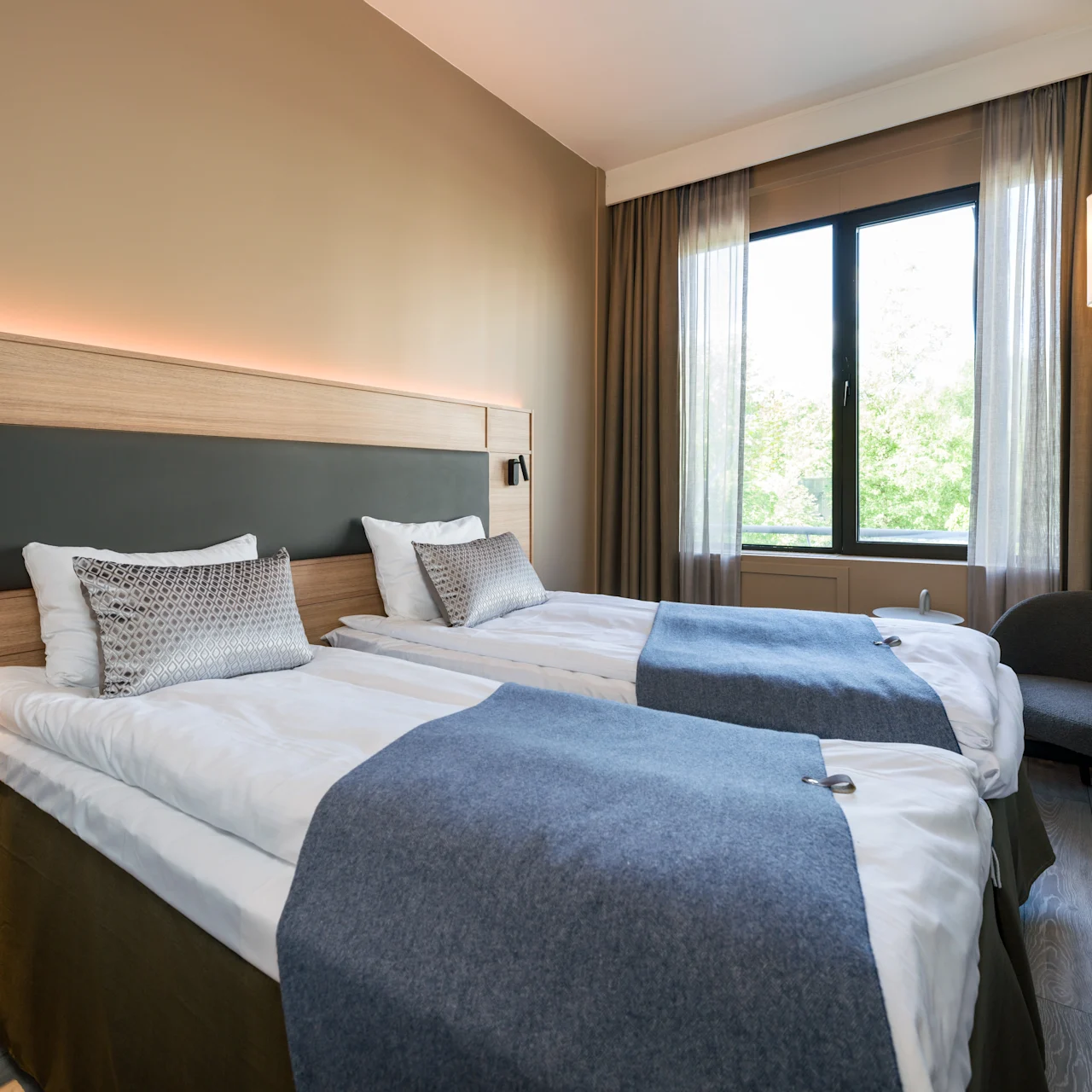 Two neatly made twin beds with gray throws are positioned side by side. A window with closed curtains illuminates the room, which features neutral walls and a modern chair.