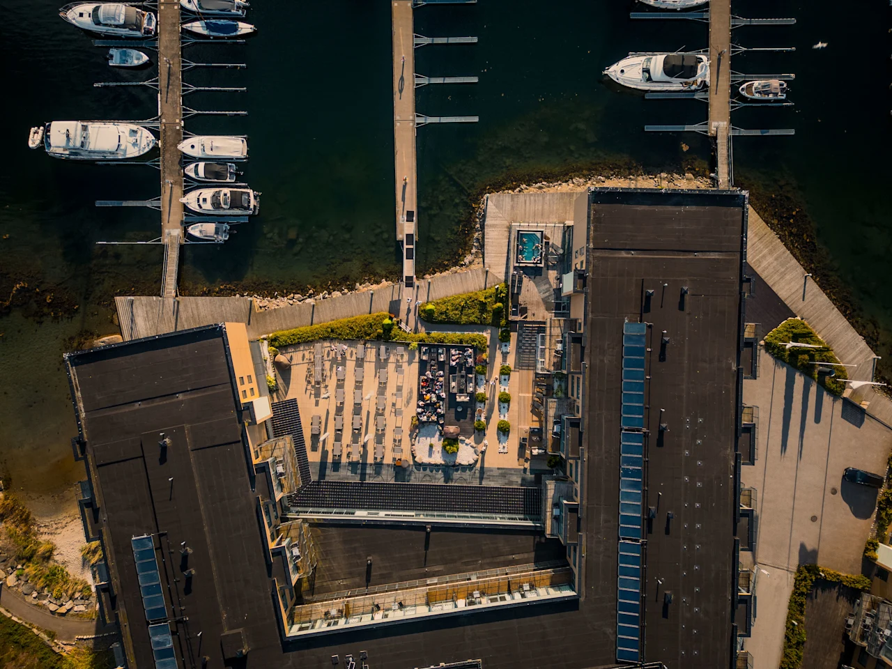 Luftfoto af en livlig marina med fortøjede både, en bygning med en livlig tagterrasse med mennesker og en forfriskende pool.