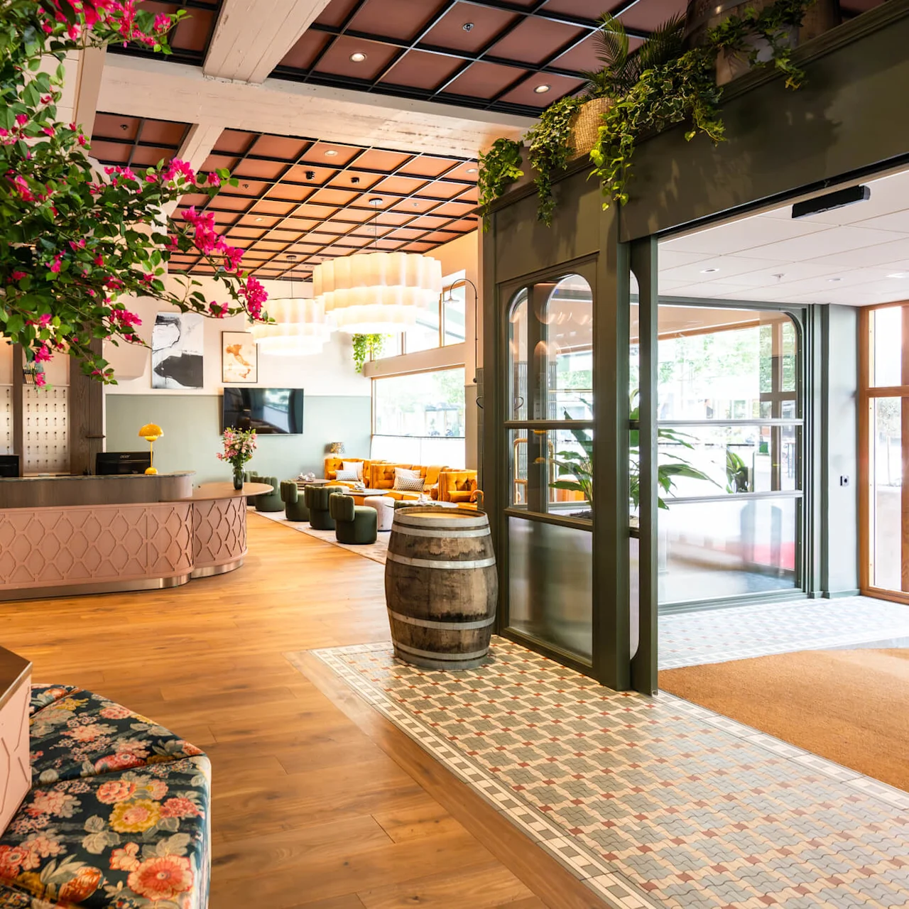 A large tree with pink flowers decorates a stylish hotel lobby featuring a wooden floor, green tiled walls, a reception desk, and cozy seating area. A decorative barrel and plants complete the setting.