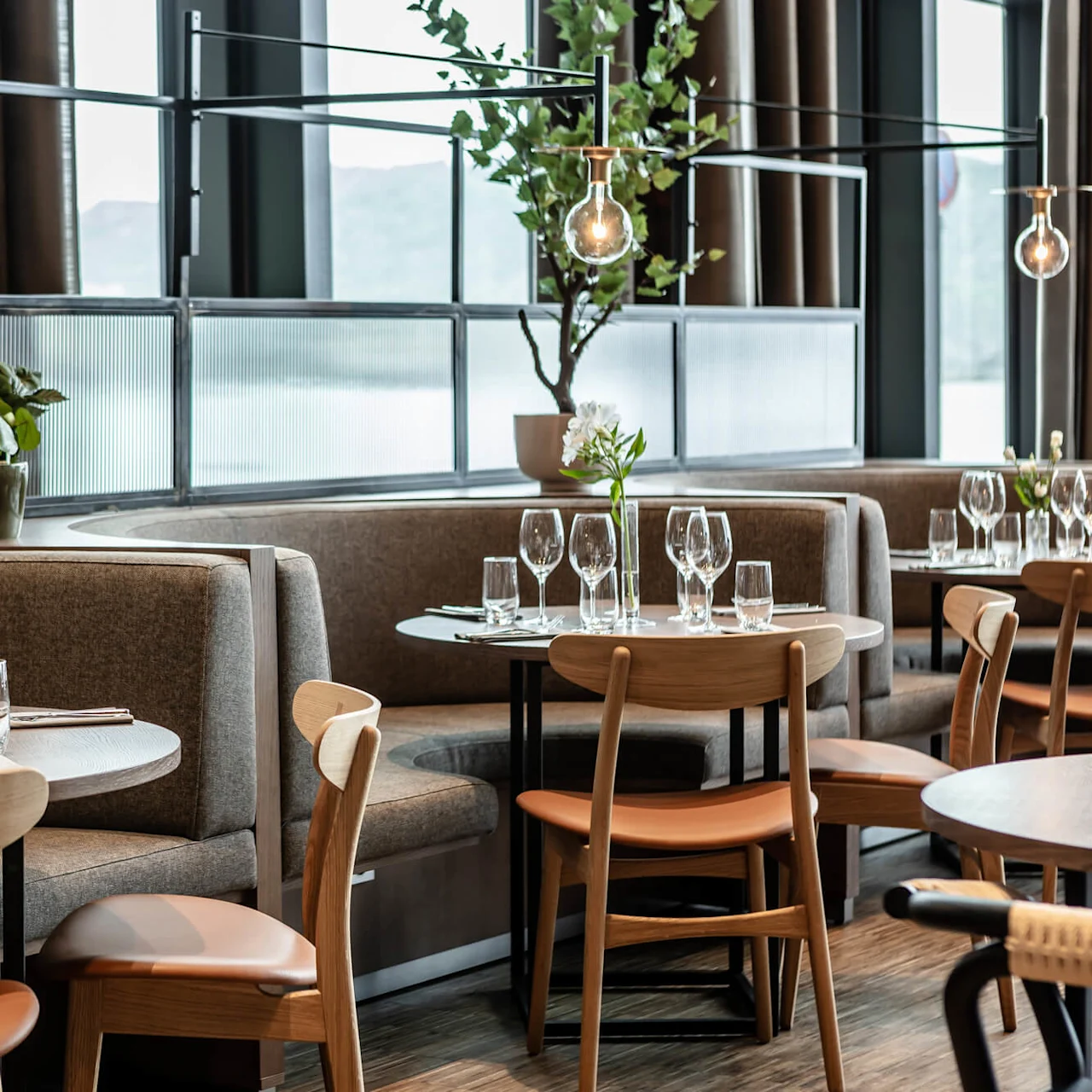 Tables are set with glassware and napkins in a modern restaurant, featuring wooden chairs, booth seating, potted plants, and pendant lighting, creating an inviting dining atmosphere.