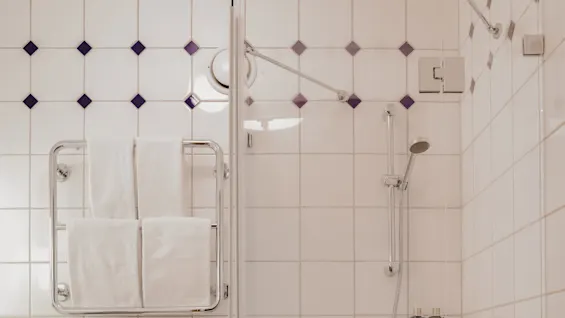 A tiled bathroom features a toilet beside a glass-enclosed shower with a mounted showerhead. Towels hang on a wall rack, and toiletries are on a small shelf.