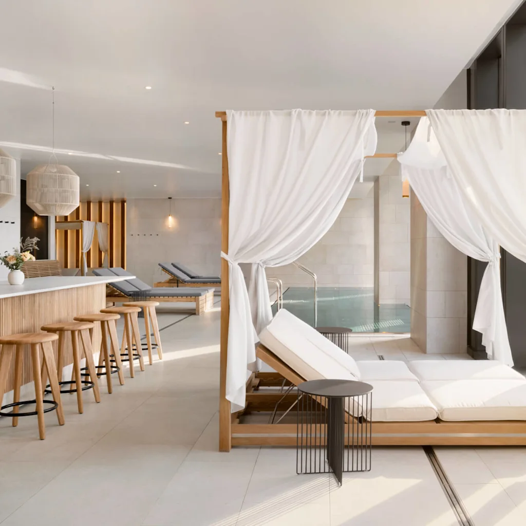 A wooden daybed with white curtains faces a modern spa environment featuring a small pool, bar area with stools, and decorative lamps. Soft lighting enhances the tranquil ambiance.