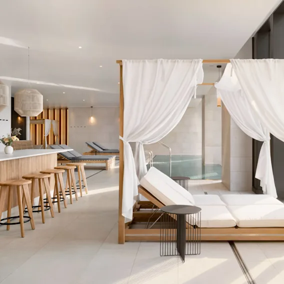 A wooden daybed with white curtains faces a modern spa environment featuring a small pool, bar area with stools, and decorative lamps. Soft lighting enhances the tranquil ambiance.