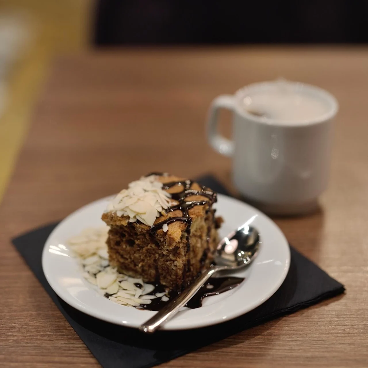 A slice of cake topped with almonds on a plate with a spoon, beside a cup of coffee on a wooden table.