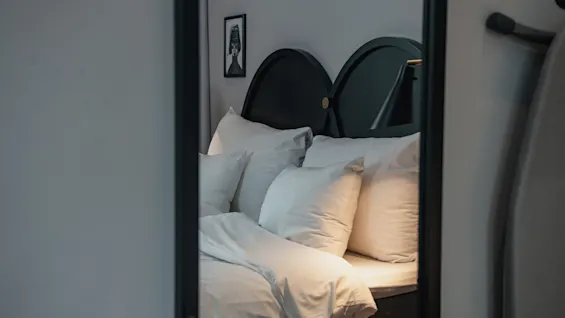 En prydligt bäddad säng med vita kuddar och lakan står i ett mjukt belyst rum, omgiven av en mörk sänggavel och dekorerad med en liten inramad bild på väggen.