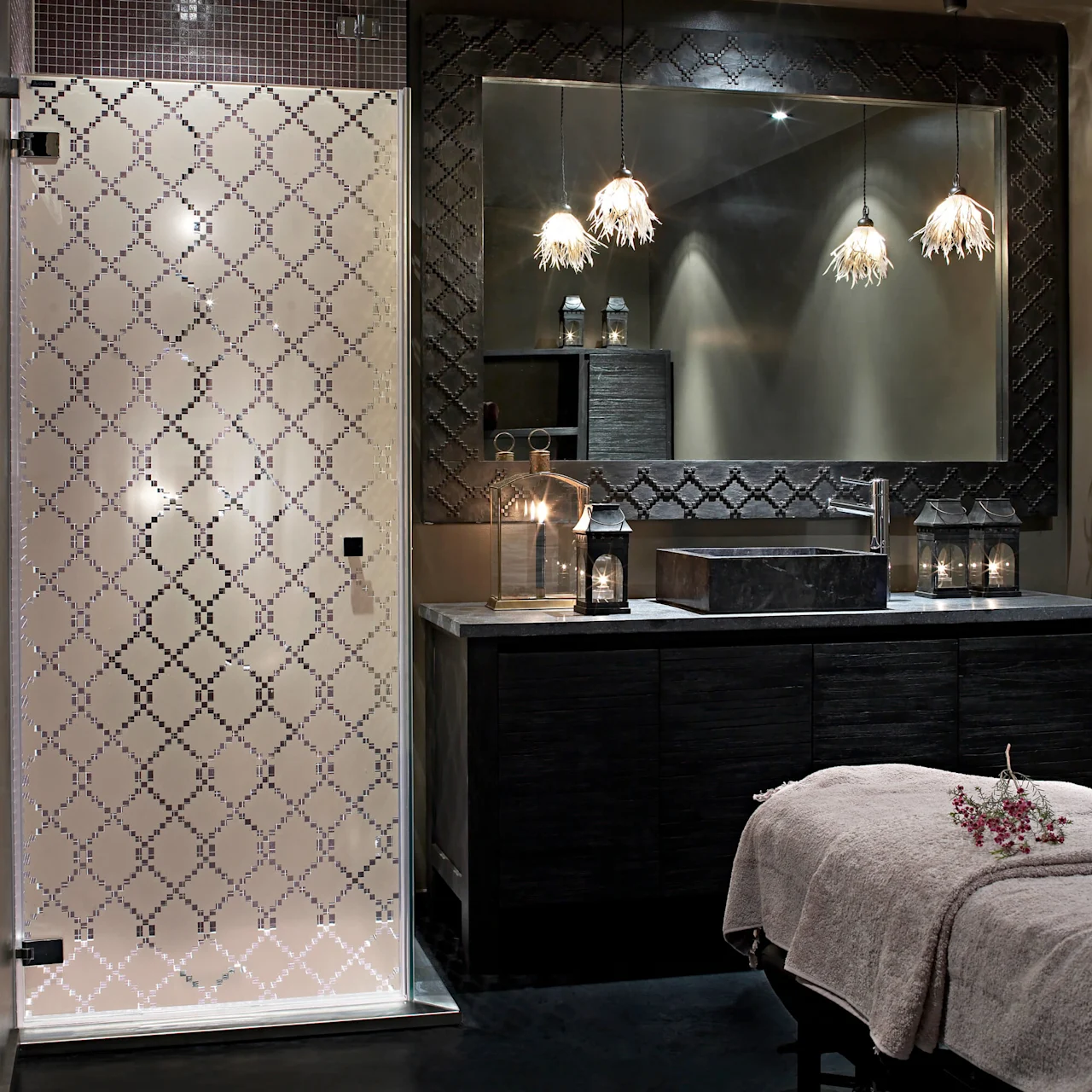 Glass shower enclosure with geometric patterns, adjacent to a dark vanity with illuminated lanterns and floral pendant lights, in a dimly lit modern bathroom setting.