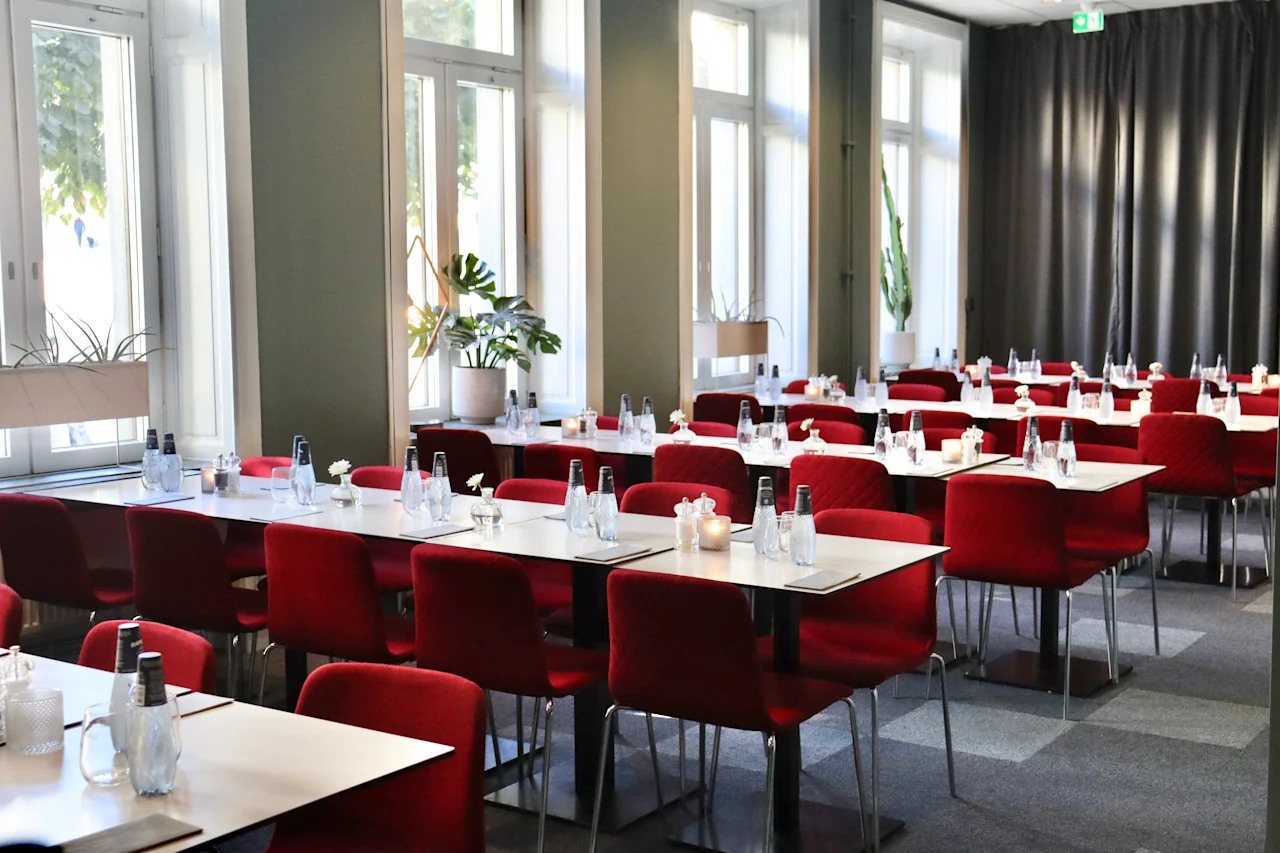 Dining tables with neatly arranged red chairs, set with water bottles and glasses, in a bright room with large windows and gray curtains.