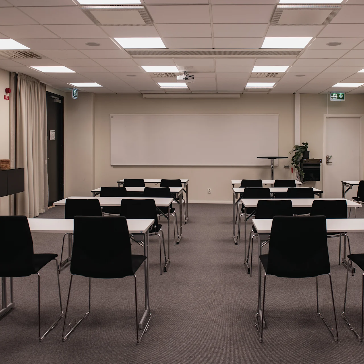 Classroom with rows of empty black chairs and white tables facing a whiteboard. A podium and emergency exit signs are visible. Ceiling lights illuminate the carpeted room.