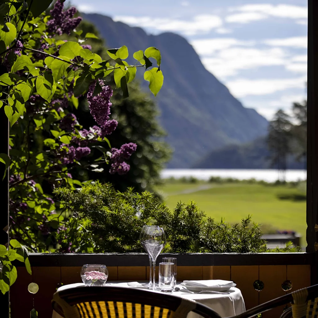 Et bord med glas er placeret på en veranda. Det har udsigt over et naturskønt landskab med frodig grønne områder, lilla blomster og fjerne bjerge ved en sø under en delvist skyet himmel.