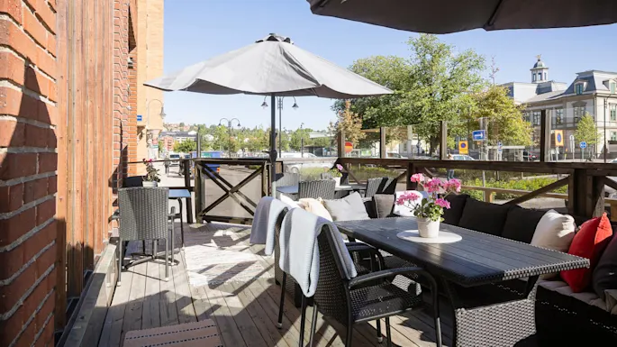 Uteområde med møbler, planter og en parasoll ved siden av en murbygning, med utsikt over en solfylt bygate.