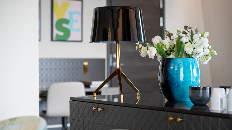 A black lamp stands on a dark patterned cabinet with gold handles. Beside it, a blue vase holds white flowers. In the background, abstract art reads "YES."