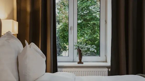 Et hyggeligt soveværelse med et vindue, der vender ud mod frodige grønne træer, en seng med hvidt sengetøj og mørke gardiner, der indrammer udsigten.